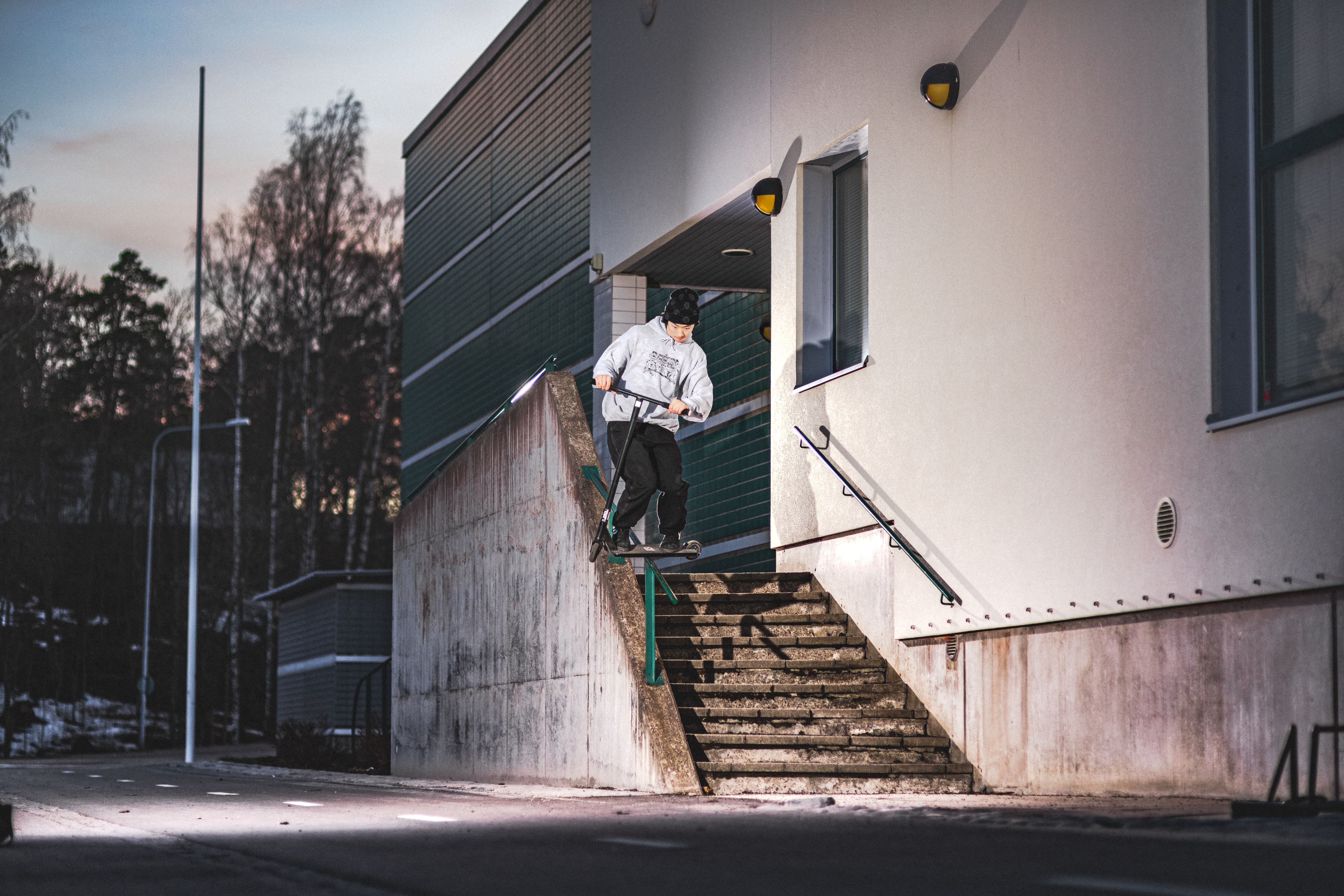 Person wearing a gray hoodie and black beanie performing a scooter grind on a handrail next to a concrete staircase at dusk.